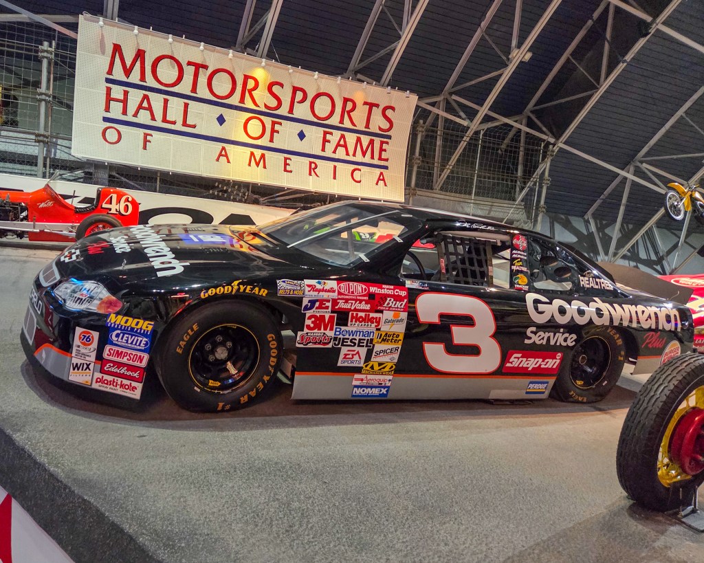 conic black NASCAR stock car number 3 with multiple sponsor decals including large white 'Goodwrench' lettering, displayed indoors in front of a 'Motorsports Hall of Fame of America' banner.