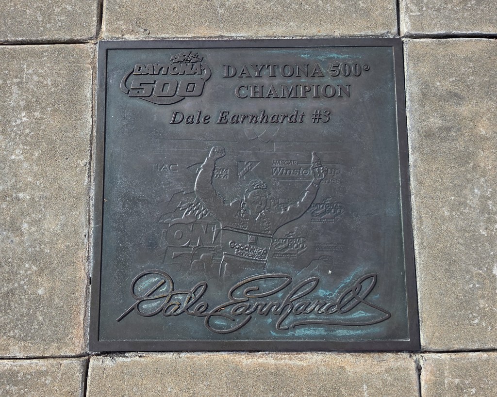 Dark bronze commemorative plaque engraved with 'Daytona 500 Champion Dale Earnhardt #3' and his signature, mounted on a wall as part of a NASCAR museum exhibit.
